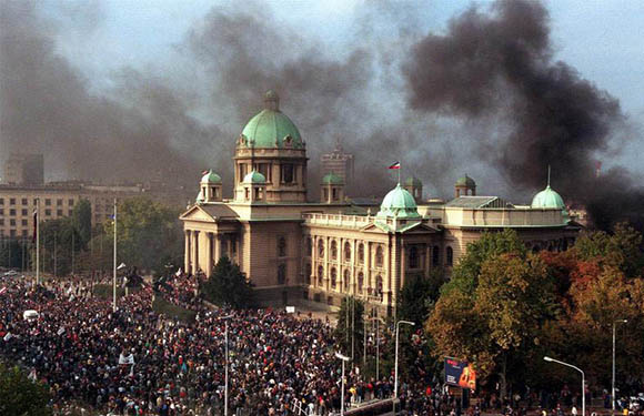 A large crowd of people gathered in front of a historic building with green domes, with smoke rising in the background.