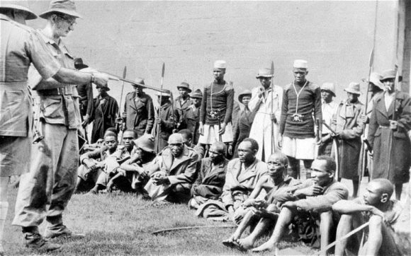 Historical photo depicts African men seated in blankets with a European soldier pointing a rifle at them, flags and soldiers behind.