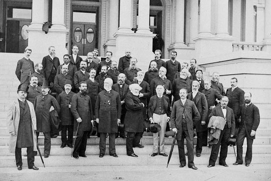 Historical photo of men in early 20th-century attire on steps of a classical building.