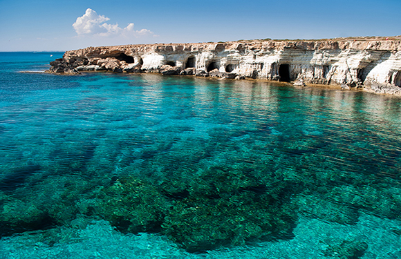 A coastal seascape with clear turquoise waters in the foreground and rocky cliffs with caves in the background under a blue sky with a small cloud.