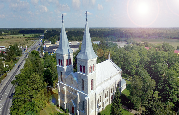 Aerial view of a large church with twin spires next to a road with surrounding greenery under a sunny sky.