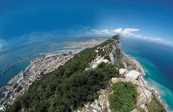 Aerial view of a coastal city with a prominent rock formation overlooking the sea and urban area, under a clear blue sky.