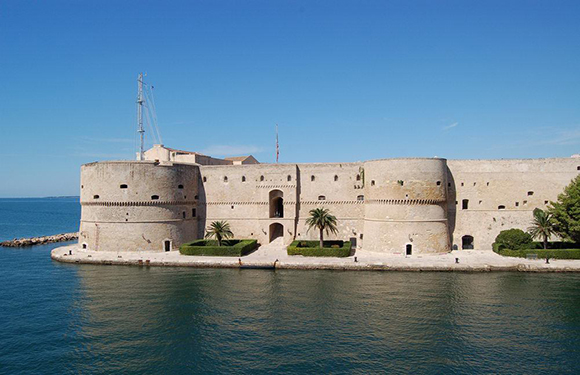 A coastal fortress with stone walls and a few trees, surrounded by calm blue waters under a clear sky.