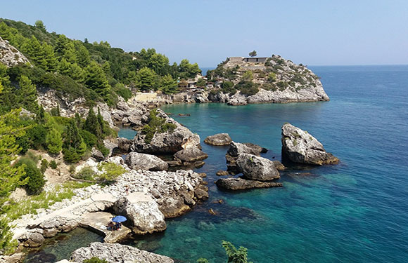 A picturesque rocky coastline with clear blue waters, boulders, cliffside greenery, and a hilltop structure.