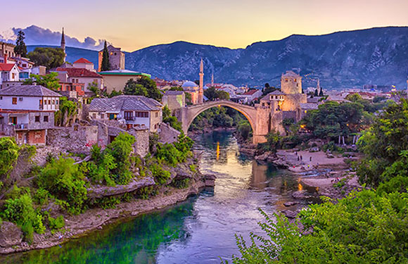 Sunset view of the historic Stari Most in Mostar, Bosnia, with old town and hills in the background.