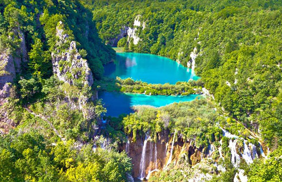 Aerial view of a turquoise lake surrounded by green forests with multiple waterfalls cascading into it.