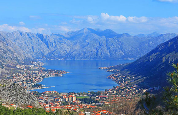 A picturesque coastal town with red-roofed buildings by a serene blue bay, framed by mountains under a semi-cloudy sky.