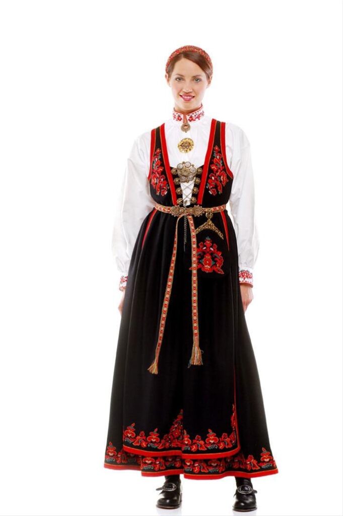 A person in traditional folk attire, featuring a black skirt, white blouse, red vest, and jewelry.