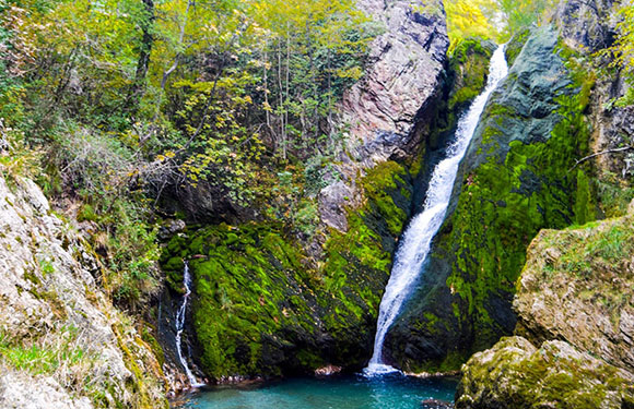 A waterfall cascading down a rocky cliff covered with green moss into a small blue pool surrounded by lush foliage.