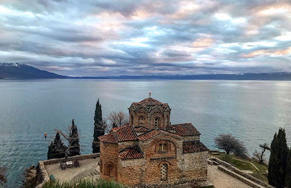 A small church with a red-tiled roof and stone walls overlooking a calm lake with mountains in the distance under a cloudy sky.