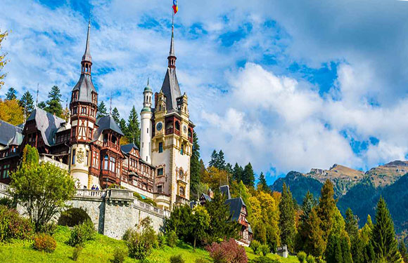 A scenic castle with many spires and intricate architecture, set on a hill amid greenery and mountains.