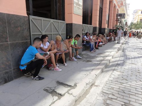 A group of people sitting on a curb along a sidewalk in a city setting, with buildings in the background and a cracked pavement in the foreground.