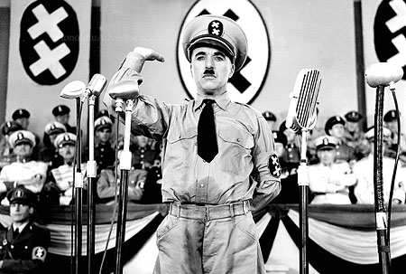 A black and white photo shows a man in a satirical military uniform, gesturing at a mic, with a backdrop of swastika-like symbols and uniformed people.