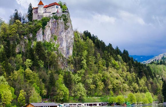 A castle sits on a cliff amidst green trees under overcast skies, with a parking lot full of vehicles below.