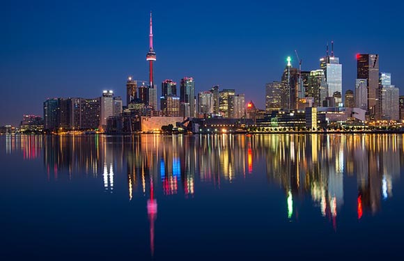 A city skyline at twilight with reflections on water, featuring a prominent tower and illuminated buildings.