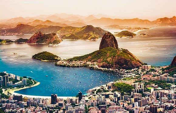 Aerial view of a coastal city with high-rise buildings near a beach, a large mountain in the background, and several islands in the bay.