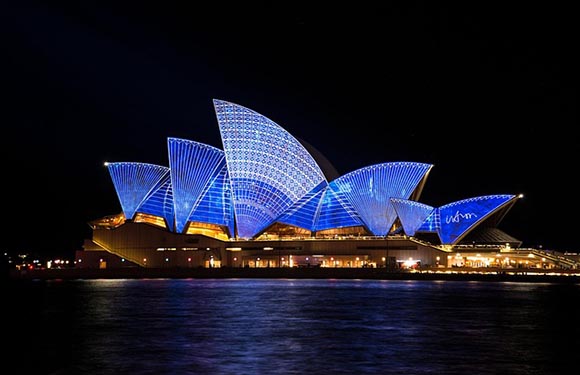 A nighttime view of the Sydney Opera House illuminated in blue light, with reflections on the water surface in the foreground.