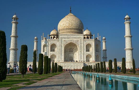 An image of the Taj Mahal with its reflection in the water, under a clear blue sky, with visitors walking around the grounds.