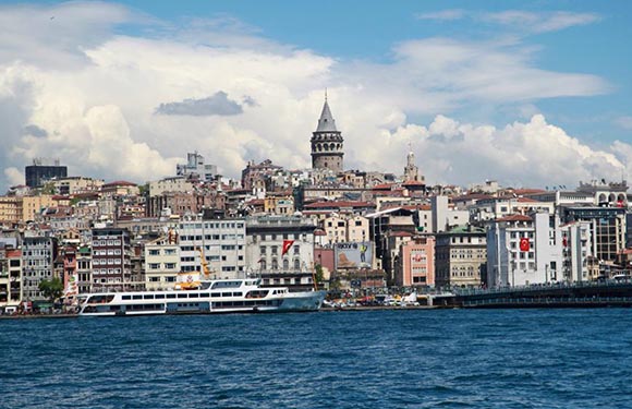 A cityscape with diverse buildings and a prominent tower, viewed from water with a visible ferry on the left.