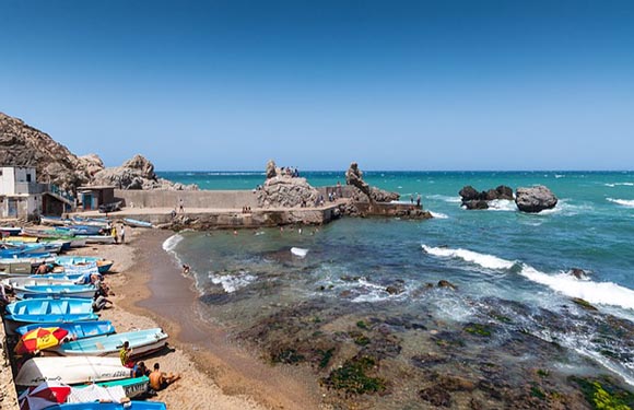 A coastal scene featuring a blue sky, turquoise sea, rocky shore, colorful beach boats, a small pier, and nearby buildings.