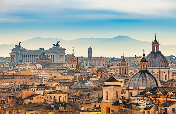 A panoramic view of a historic cityscape with domed buildings and distant mountains under a hazy sky.