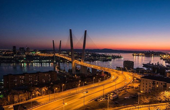 Aerial dusk view of a city with a lit cable-stayed bridge over water, glowing streets, and buildings against twilight sky.