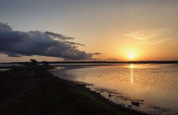 A sunset view over a calm body of water with reflections of the sun on the surface, clouds in the sky, and land visible on the left side.