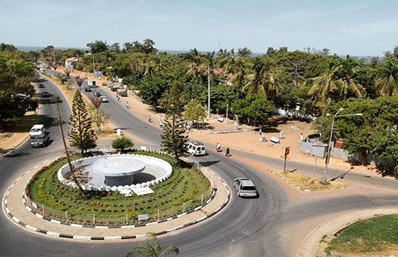 Aerial view of a roundabout with vehicles, surrounded by greenery and palm trees, under a clear sky.