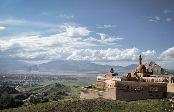 A historic castle with a dome and towers sits on a hill, overlooking a vast landscape with distant mountains under a partly cloudy sky.