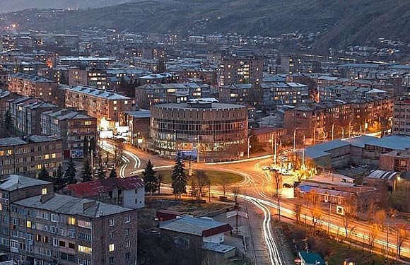 A nighttime cityscape with illuminated streets and buildings, nestled in a valley with mountains in the background.