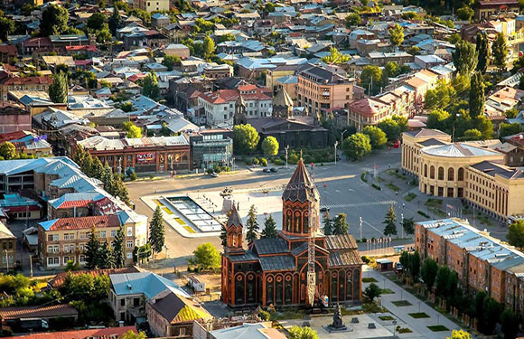 Aerial shot of a city with a tall, brick church, various buildings, greenery, bathed in warm sunlight.