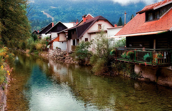 A scenic view of houses along a calm river with greenery and mountains in the background.