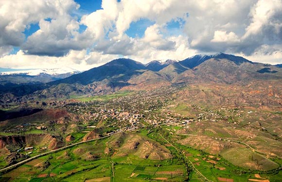 Aerial view shows green fields, a town in a valley, surrounded by brown mountains under a cloudy sky.