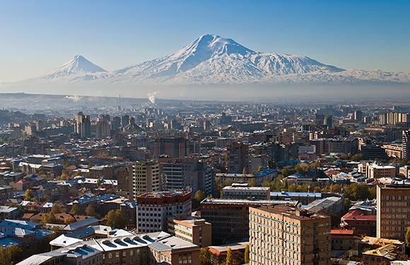 A cityscape with numerous buildings under a clear sky, with a large snow-capped mountain in the background.