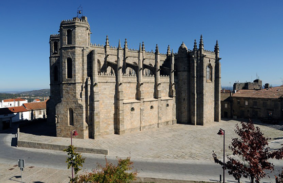 A Gothic stone church with a square bell tower is set on a paved area with distant buildings, trees, and a clear blue sky backdrop.