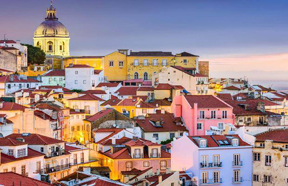 A European city at dusk with terracotta-roofed buildings, a church dome, and a pastel sky.