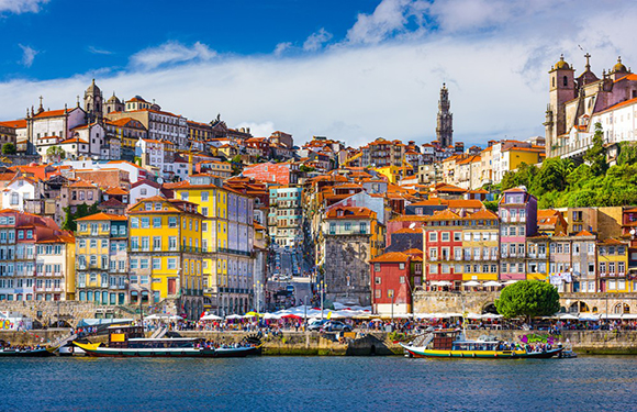 A vibrant cityscape with historic, colorful buildings on a hillside, overlooking a river with boats under a clear sky.