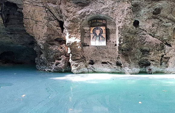 A serene water body in front of a rocky cave with an icon of a religious figure displayed in a niche.