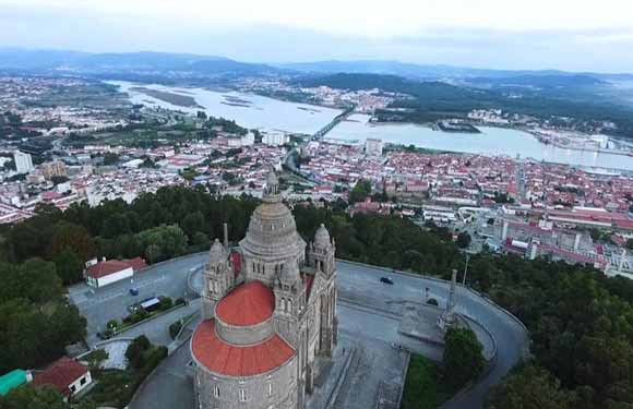 Aerial view of a large church with a dome and twin towers, surrounded by roads and trees, with a panoramic backdrop of a town divided by a river.