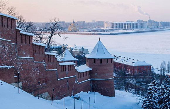 A snowy winter scene features a brick fortress with towers, overlooking a river and cityscape under a dusky sky.