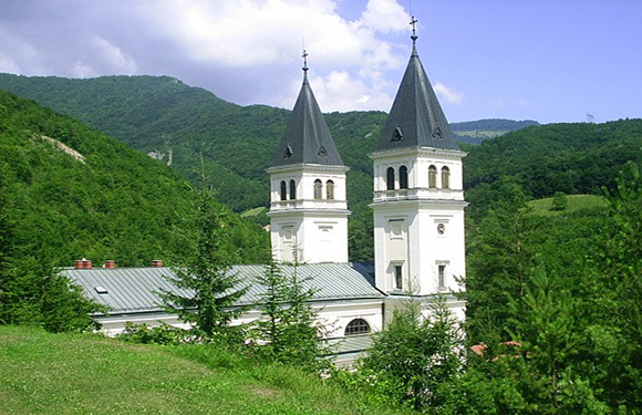 A white church with two prominent spires nestled in a lush green valley with forested hills in the background.