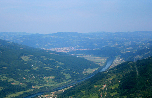 A scenic view of a valley with a river running through it, surrounded by green hills and a few scattered buildings, under a hazy blue sky.