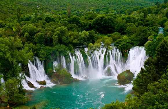 A scenic view of multiple waterfalls cascading into a turquoise pool surrounded by lush green foliage.