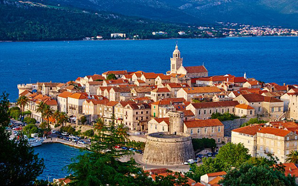 A scenic coastal town with historic architecture, bell tower, red roofs, encircled by water and hills.