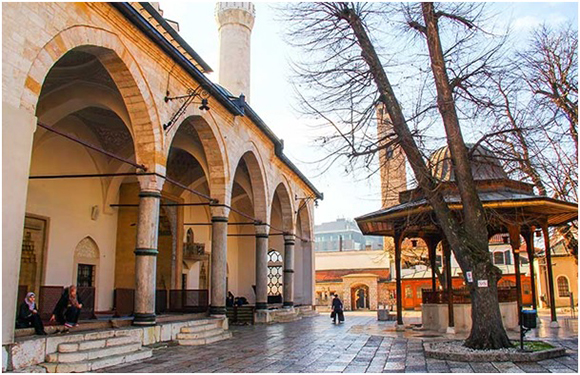 An image of a mosque with an arched outdoor gallery, a minaret, and a small fountain or pavilion in a courtyard with leafless trees and a few people.