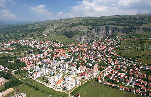 Aerial view of a town with dense housing and several larger buildings, surrounded by green fields and hills in the background.