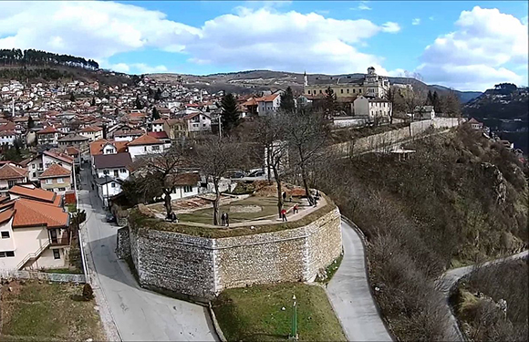 Aerial view of a historic town with dense buildings, a fortified wall in the foreground, and hills in the background under a cloudy sky.