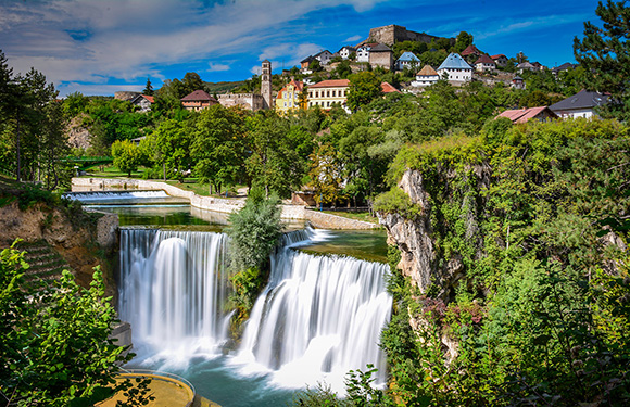 A picturesque waterfall with many cascades, set amid greenery, with a town and hilltop fortress under a semi-cloudy sky.