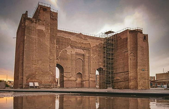 A historical brick structure with arches and scaffolding against a cloudy sky, reflected in a body of water in the foreground.