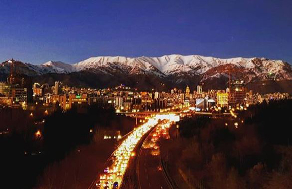 A cityscape at night with lit buildings and streets, a bustling central road, and snowy mountains under a twilight sky.